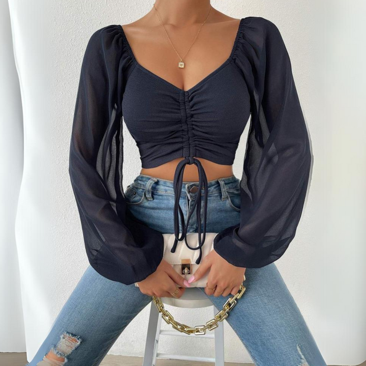 A woman wearing a black long-sleeve sheer crop top with a flirty neckline, blue ripped jeans, and a gold necklace sits on a stool holding a white handbag with a chunky gold chain. The background is plain and white.
