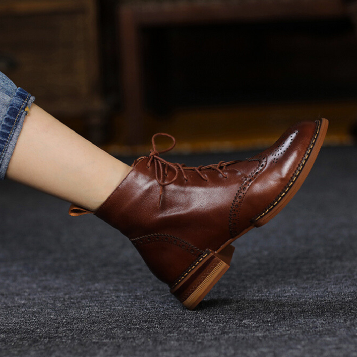 A person wearing dark blue rolled-up jeans and genuine leather ankle boots with decorative stitching and a low heel, standing on a dark carpeted floor—an effortless look for the urban trendsetter.