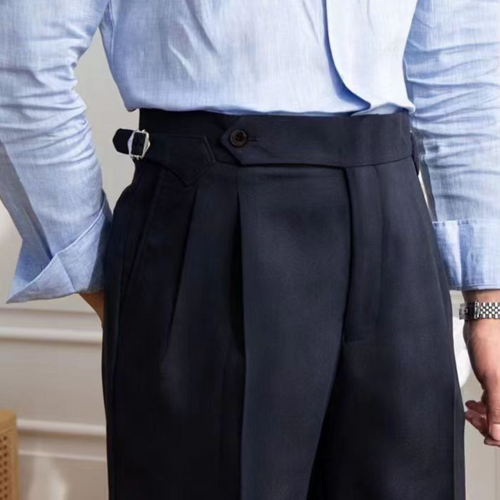 A person wearing a tailored fit light blue dress shirt tucked into high-waisted navy trousers with side adjusters, pleats, and a buttoned waistband. The left hand rests by the side, a watch visible on the wrist for an old money aesthetic.