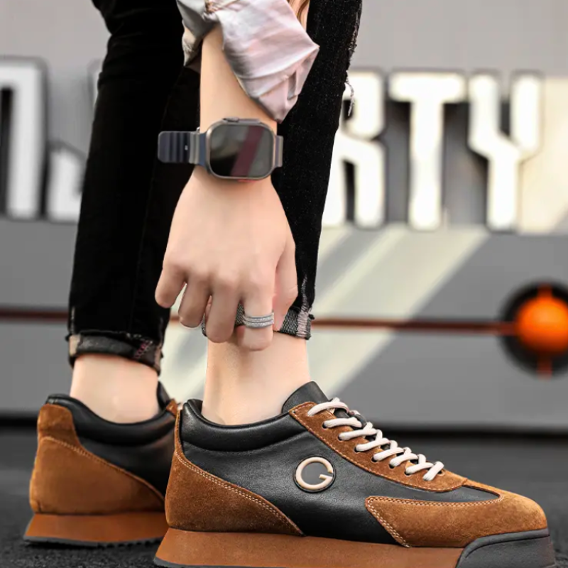 A person wearing stylish brown and black luxe leather suede sneakers with a G logo bends down, revealing a smartwatch and rings on their hand. The blurred, modern wall with bold geometric patterns adds an urban professional vibe.