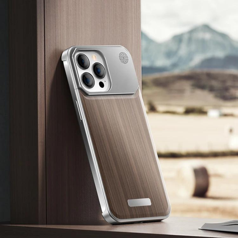 A smartphone with a stylish wood phone case and metallic accents rests upright against a wooden surface. Mountains and fields are blurred in the background, viewed through a window.