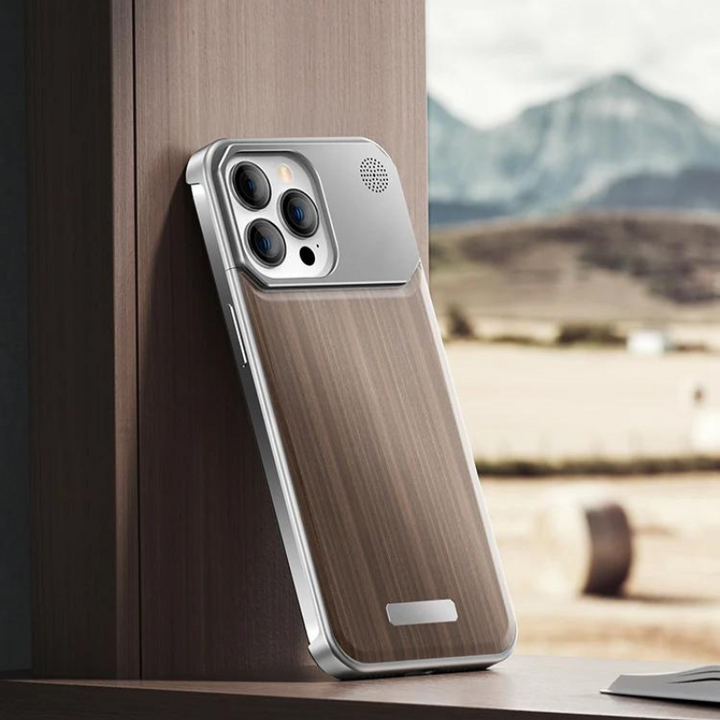 A smartphone with a stylish wood phone case and metallic accents rests upright against a wooden surface. Mountains and fields are blurred in the background, viewed through a window.