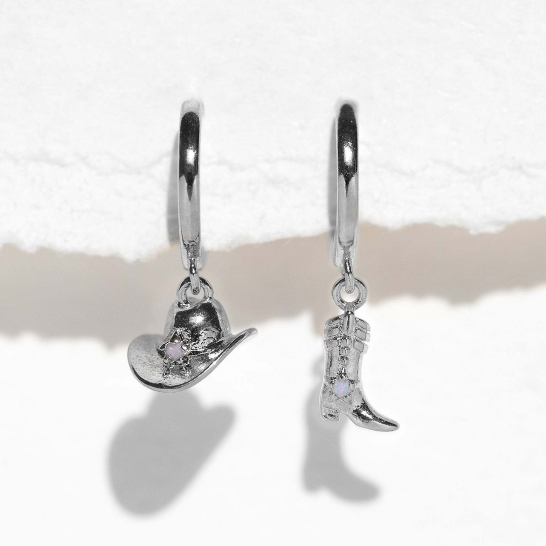 Two silver hoop statement earrings hang against a white textured background; one earring features a cowboy hat charm and the other a cowboy boot charm, each adorned with a small clear gemstone.