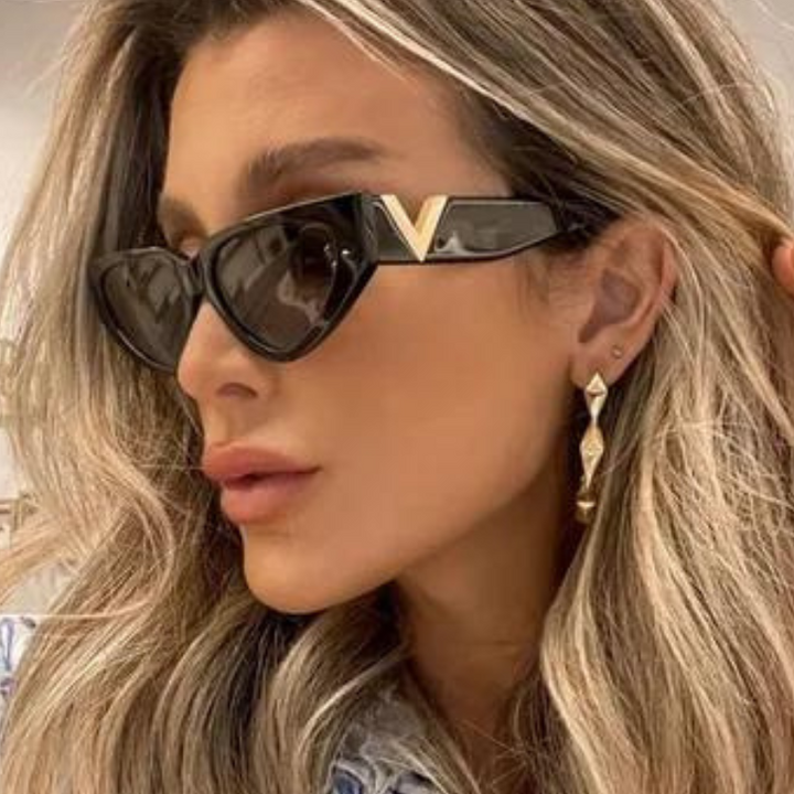 A woman with wavy blonde hair wears bold frame sunglasses with gold V accents and large gold earrings. She looks to the side, showing off her chic accessories, UV400 protection, and styled hair.