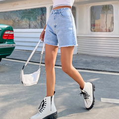 A person wearing a white crop top, high-waist denim shorts, white combat boots, and carrying a white handbag is walking outdoors near a green car and a building with large windows.