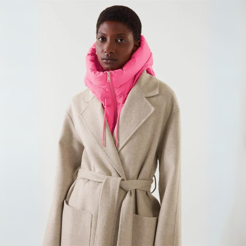 A person wearing a beige belted coat with a bright pink puffer vest featuring a hooded design stands against a plain light background.