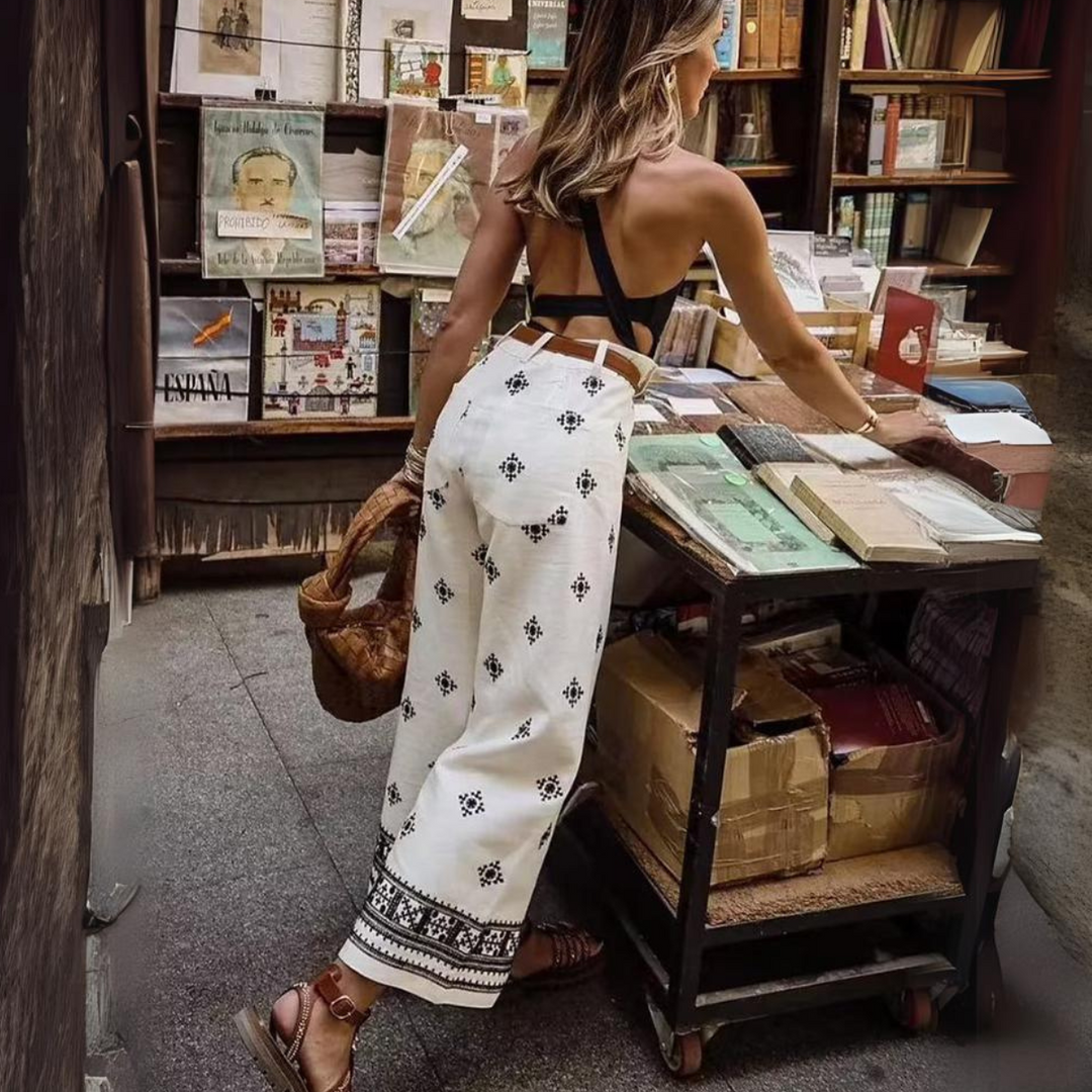 A woman in a backless black top and boho chic pants browses an outdoor bookstall filled with books and magazines. She holds a woven bag and stands on a stone walkway next to the display.