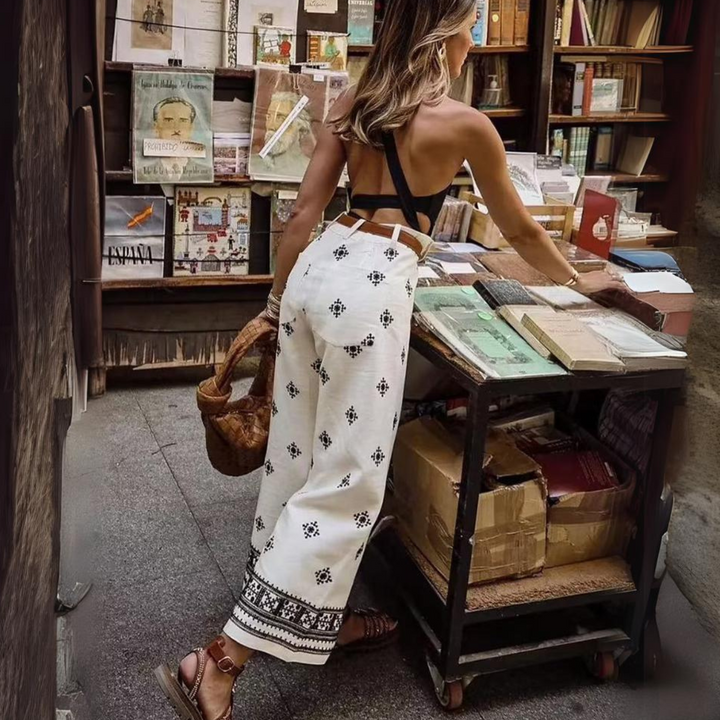 A woman in a backless black top and boho chic pants browses an outdoor bookstall filled with books and magazines. She holds a woven bag and stands on a stone walkway next to the display.