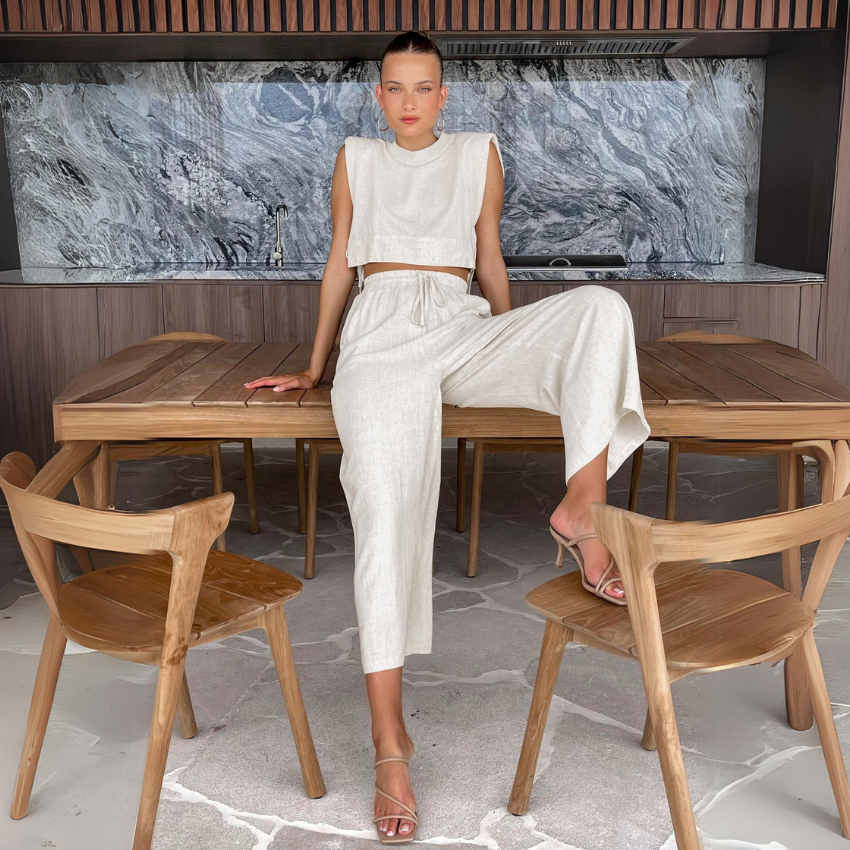 A woman in a sleeveless cropped top and matching wide-leg pants sits on a wooden dining table with one leg up, surrounded by wooden chairs, with a modern marble backsplash and wood cabinetry in the background.