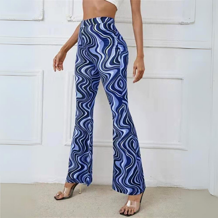A person wearing trendy statement pants—high-waisted, blue and white wavy patterned with clear high-heeled sandals—stands against a white-paneled wall. Only the lower half of the body is visible.