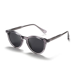 Fashionable eyewear featuring round frame sunglasses with dark lenses and translucent gray frames, displayed on a white background. The arms are partially folded, and the glasses are angled slightly to the side.