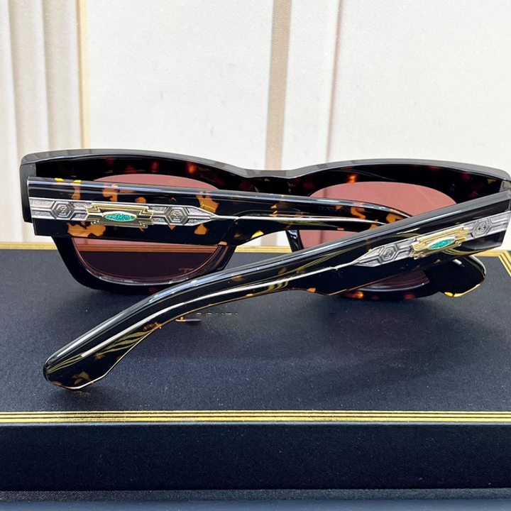 A pair of vintage-inspired tortoiseshell sunglasses with silver and turquoise detailing on the arms rests on a black box with gold trim, against a light-colored, striped background.