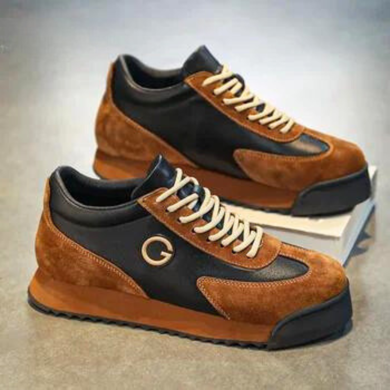 A pair of sleek sneakers with brown suede and black luxe leather panels, cream-colored laces, and a gold G logo on the side, placed on a gray surface with one shoe propped atop a white box.