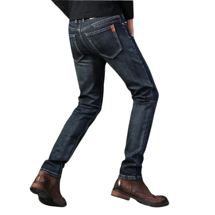 A person wearing dark blue slim-fit jeans crafted from durable fabric and brown leather boots, photographed from the back against a white background.