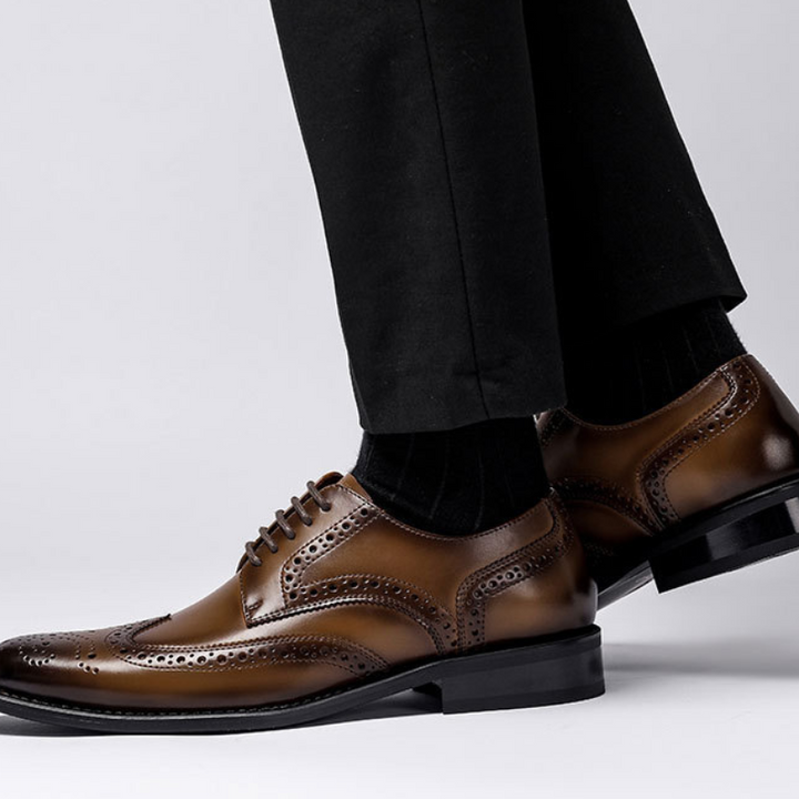 A person wearing polished brown premium leather brogue dress shoes with a cushioned insole and slip-resistant rubber sole, paired with black socks and black dress pants, stands against a neutral background.