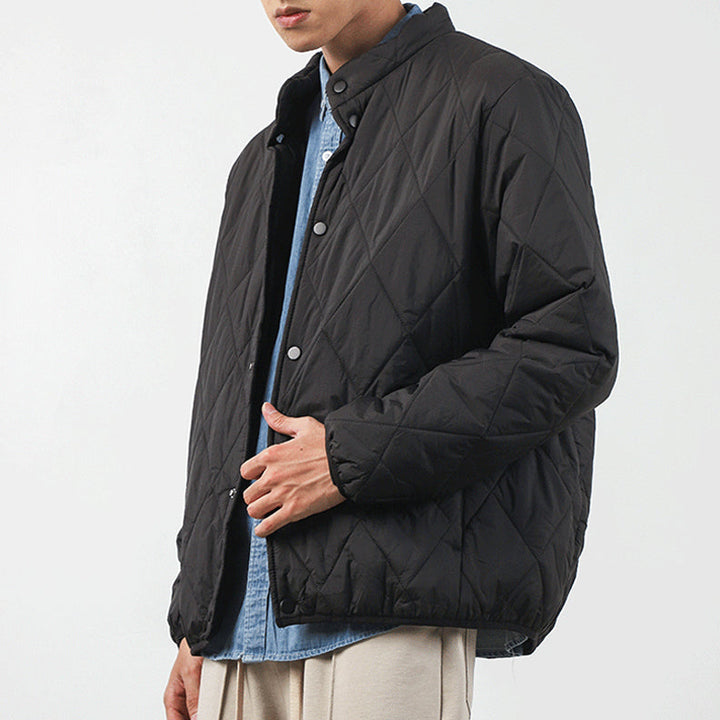 A person wearing a black, lightweight insulating jacket with a diamond-stitched pattern over a light blue button-up shirt and light-colored pants, standing against a plain white background. Their face is partially out of frame.