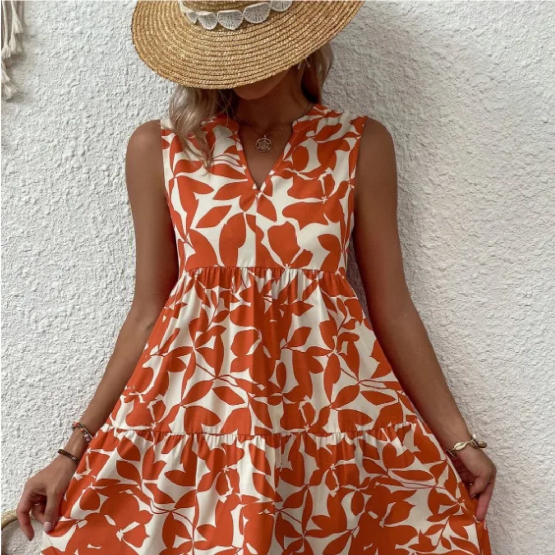 A woman in a sleeveless, orange and white leaf-pattern dress made of lightweight breathable fabric and a wide-brimmed straw hat stands against a textured white wall, holding the sides of her dress. Her face is mostly covered by the hat.
