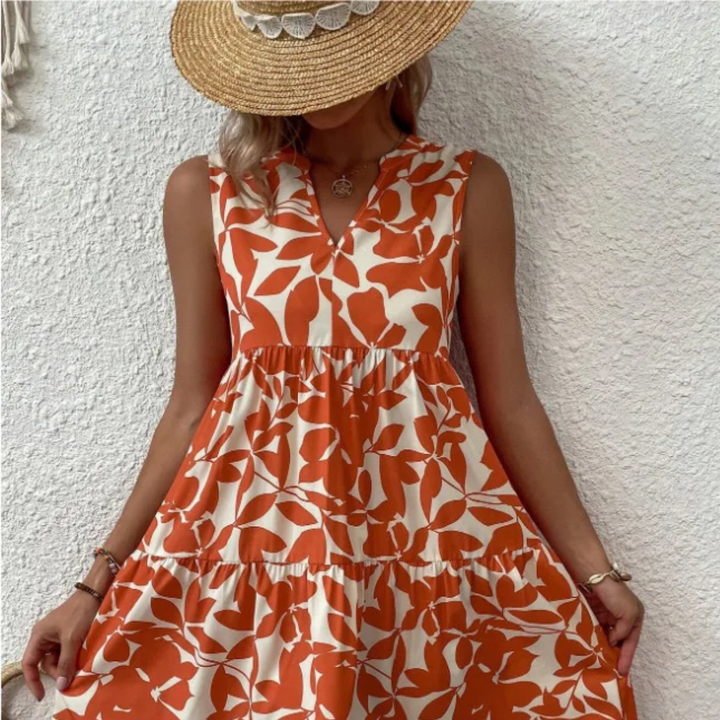 A woman in a sleeveless, orange and white leaf-pattern dress made of lightweight breathable fabric and a wide-brimmed straw hat stands against a textured white wall, holding the sides of her dress. Her face is mostly covered by the hat.