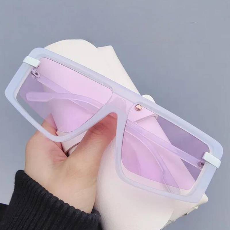 A hand holds a pair of oversized, rectangular sunglasses with light purple-tinted UV400 lenses and white polycarbonate frames against a plain grey background, offering both style and sun protection.