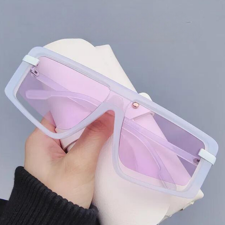 A hand holds a pair of oversized, rectangular sunglasses with light purple-tinted UV400 lenses and white polycarbonate frames against a plain grey background, offering both style and sun protection.