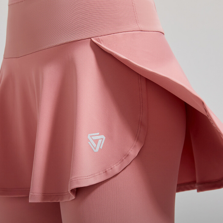 A close-up of a person wearing a pink athletic skort with high-waisted support, featuring a flared skirt over fitted shorts. A white geometric logo is visible on the skirt. The background is a plain light color.