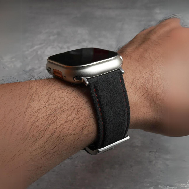 A close-up of a person’s wrist wearing a smart watch with a dark suede strap, red stitching, and stainless steel buckle, set against a textured gray background.