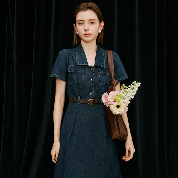 A woman wearing a belted denim dress, perfect for summer wear, stands in front of a black curtain, holding a brown shoulder bag filled with pink and white flowers. She has straight hair and a neutral expression.