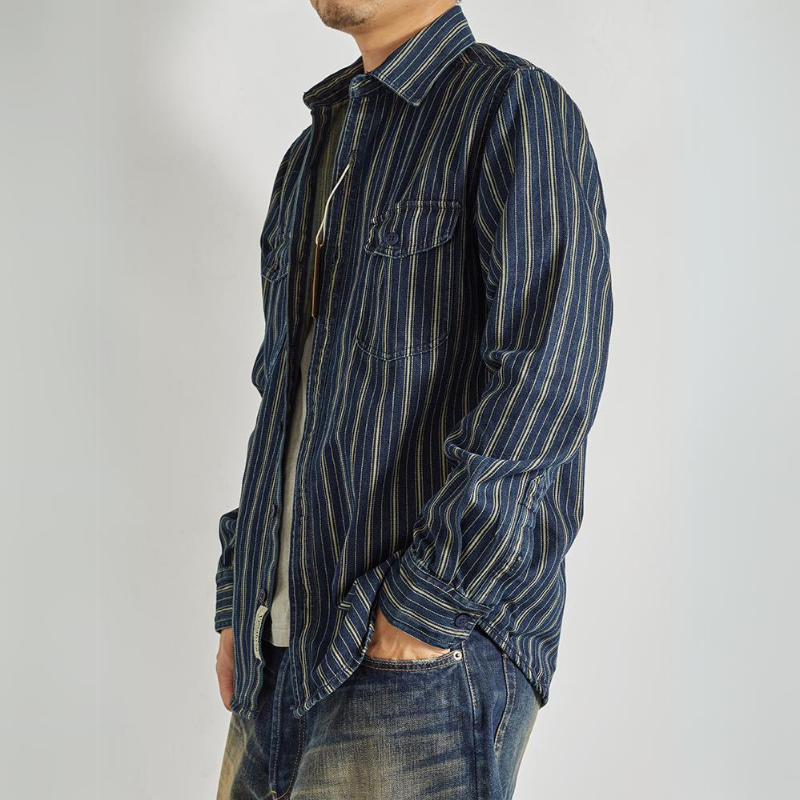 A person wearing a relaxed-fit button-down with vertical stripes over a light-colored undershirt and faded blue jeans, standing with one hand in their pocket against a plain light background.