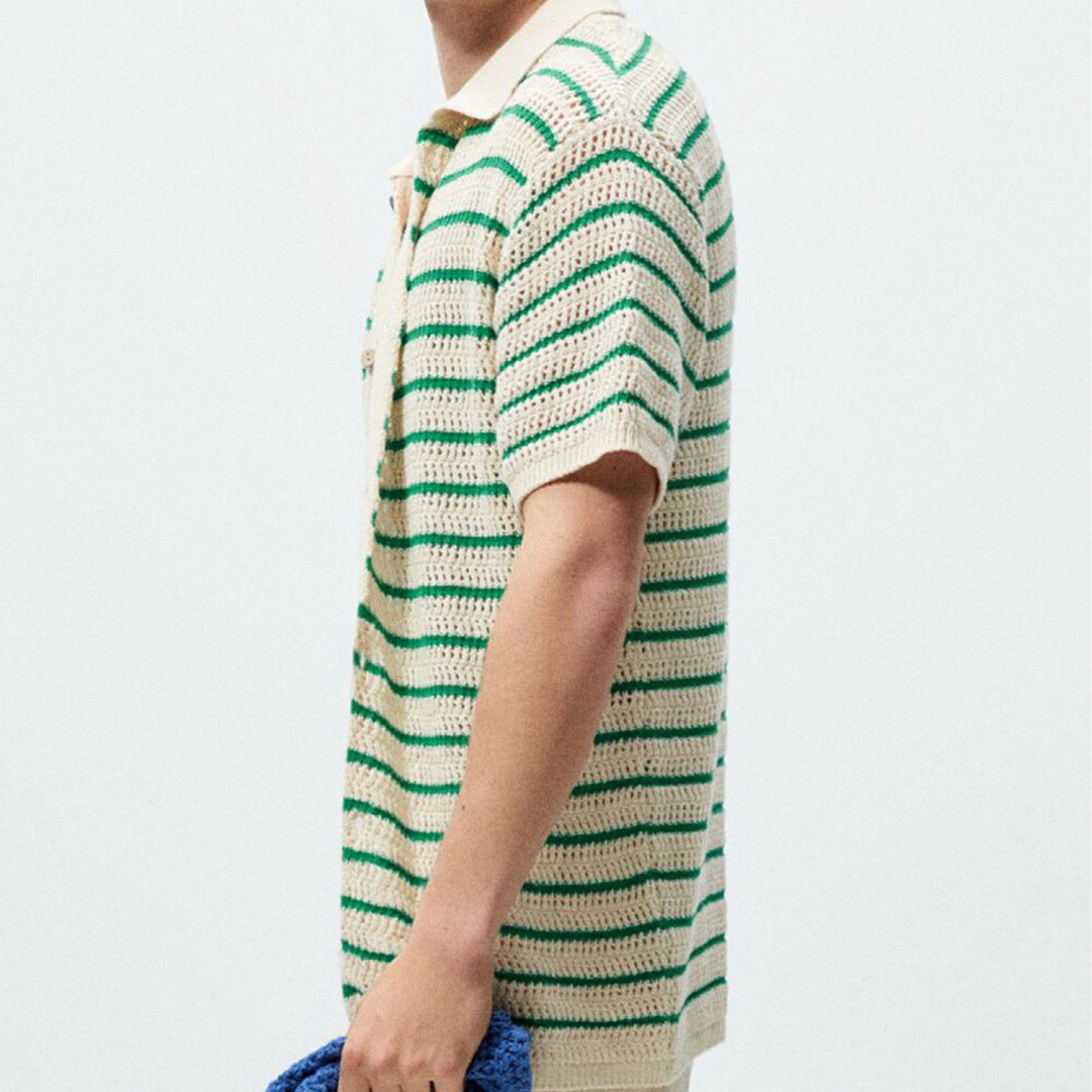 A person wearing a short-sleeve, cream-colored open-knit design shirt with green horizontal stripes stands sideways, holding a textured blue item in their hand against a plain light background.