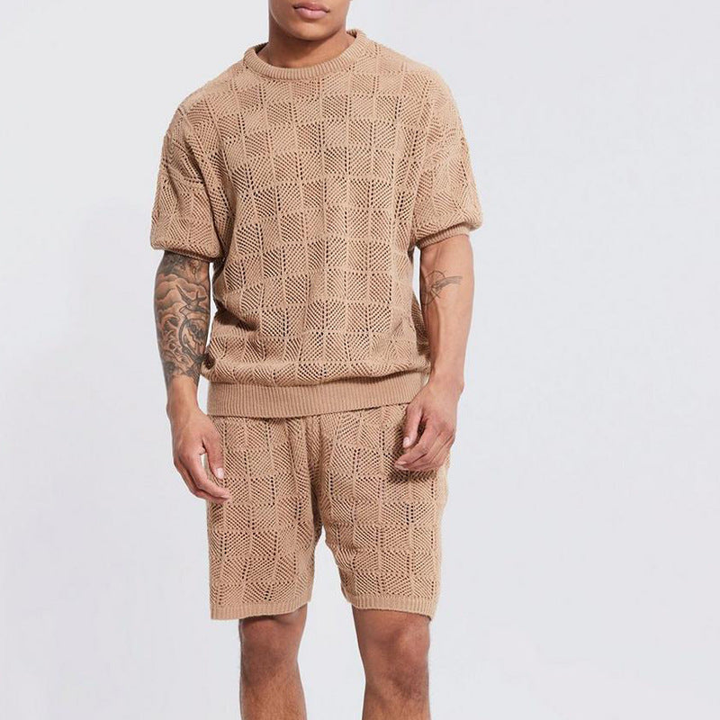 A man stands against a plain background wearing a men's 2 piece set: a matching knit beige short-sleeve top and shorts with a textured checkered pattern. His right arm is tattooed, and his face is partially out of frame.