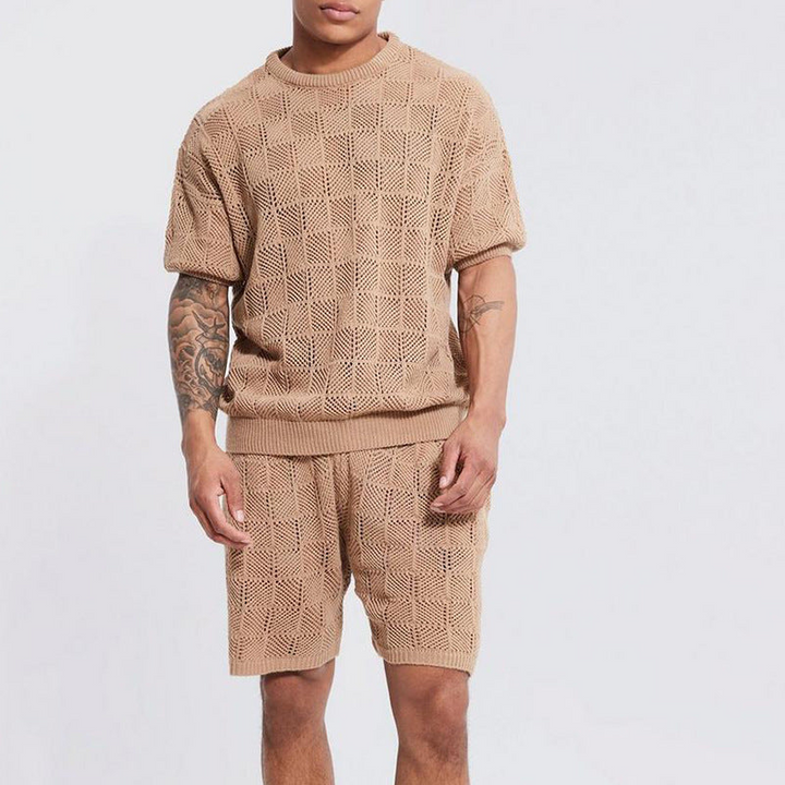 A man stands against a plain background wearing a men's 2 piece set: a matching knit beige short-sleeve top and shorts with a textured checkered pattern. His right arm is tattooed, and his face is partially out of frame.