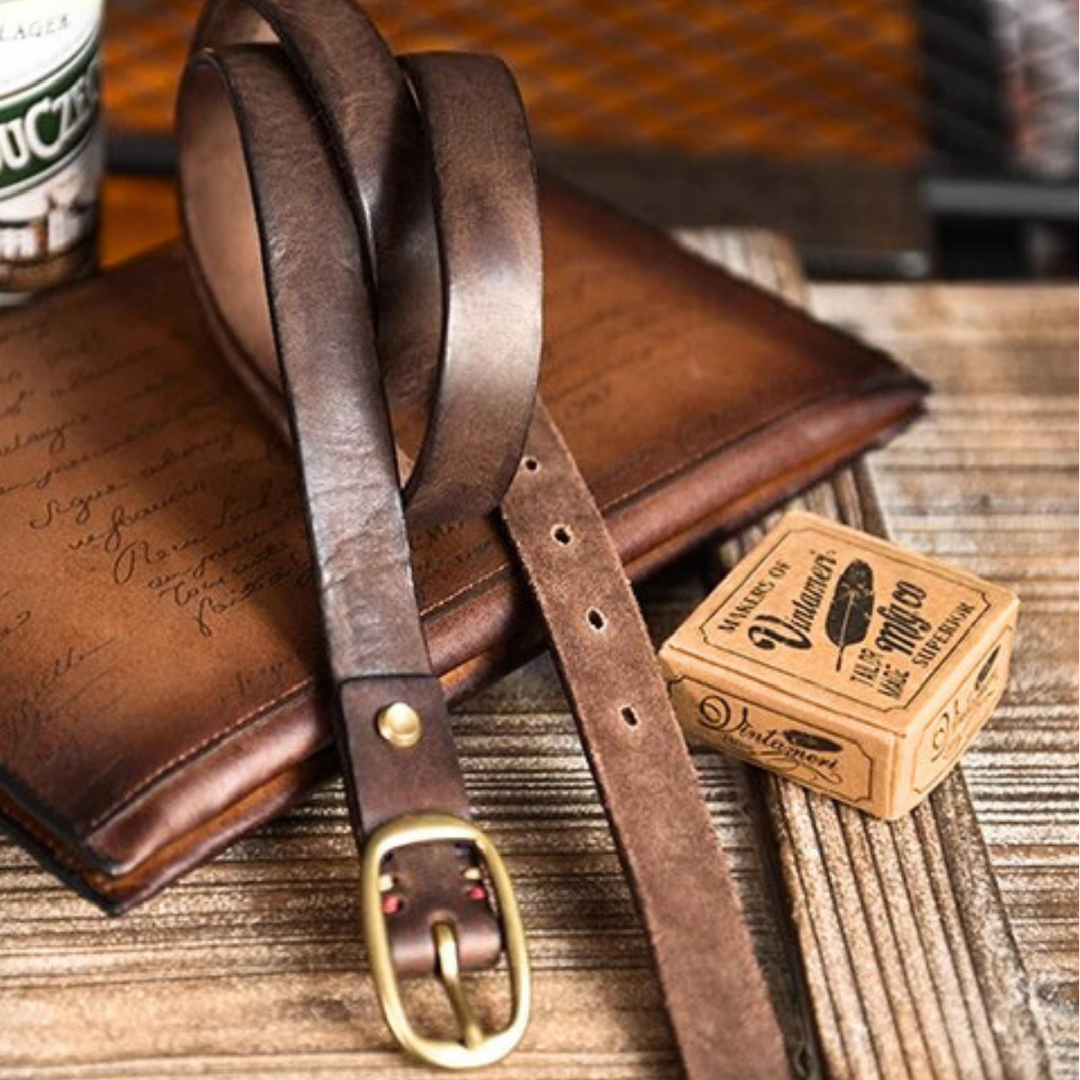 A brown genuine leather belt with a brass buckle rests on a vintage leather-bound notebook next to a small box of leather conditioner, all arranged on a wooden surface exuding classic Southern style.