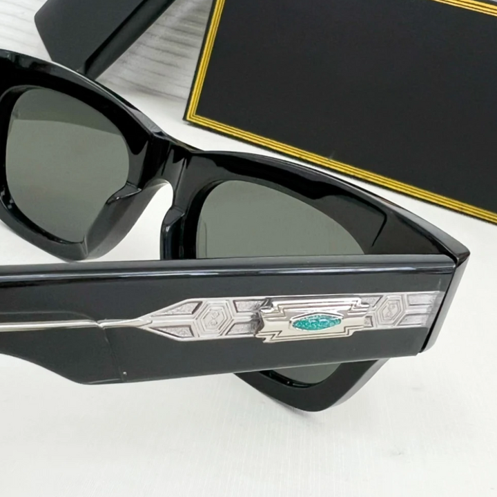 Close-up of retro sunglasses with an intricate silver and turquoise geometric design on the temple, resting on a white surface with a black and gold case in the background.