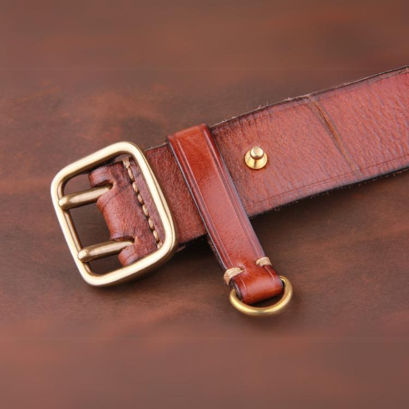 A close-up of a vintage leather belt crafted from premium leather, featuring a gold buckle and gold ring, lying on a brown surface. The leather’s visible texture and stitching highlight its quality craftsmanship.