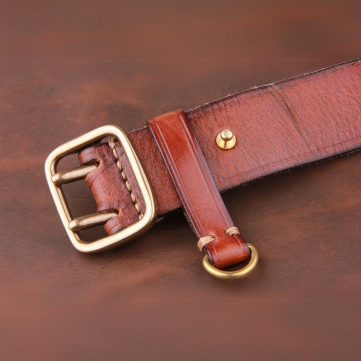A close-up of a vintage leather belt crafted from premium leather, featuring a gold buckle and gold ring, lying on a brown surface. The leather’s visible texture and stitching highlight its quality craftsmanship.