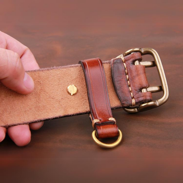 A hand holding a vintage leather belt, showing the suede underside, brass buckle, and metal ring on a wooden surface.