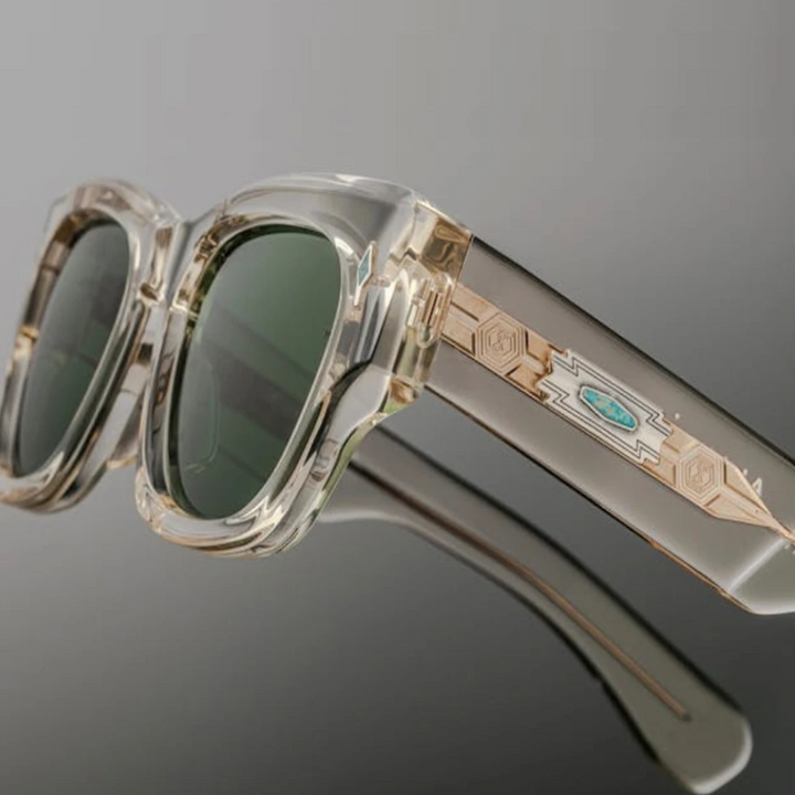 Clear-framed retro sunglasses with green lenses and decorative gold and turquoise geometric detailing on the arms, offering UV400 protection, displayed on a reflective gray surface.