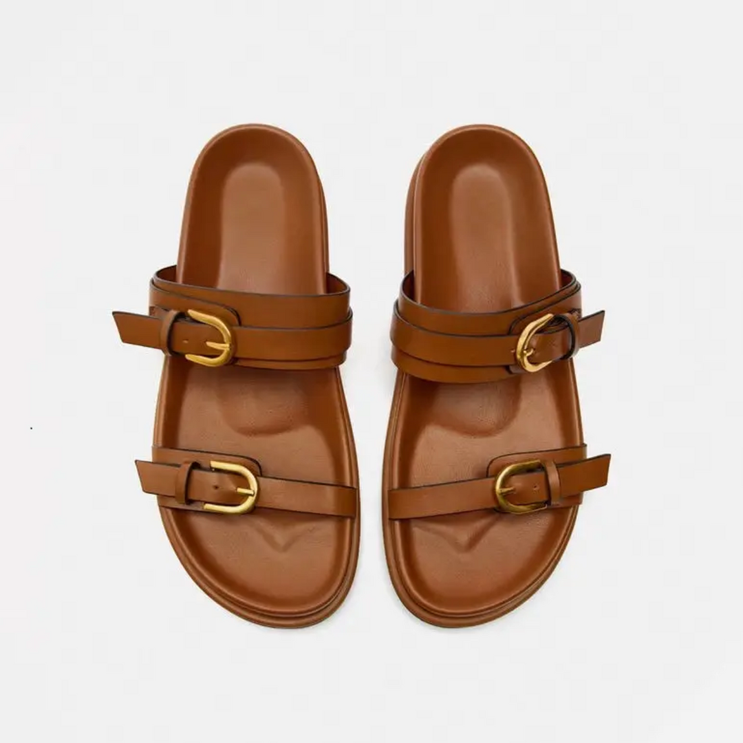 A pair of brown leather slide sandals with a leather upper, two straps, and gold buckles on each sandal, displayed on a plain white background.