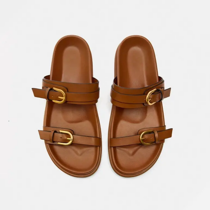 A pair of brown leather slide sandals with a leather upper, two straps, and gold buckles on each sandal, displayed on a plain white background.