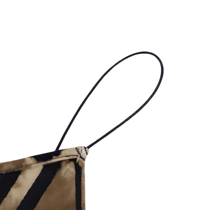 A close-up of a fabric face mask with a tiger stripe pattern, reminiscent of an animal print dress, showing one ear loop and part of the mask’s corner against a plain white background.