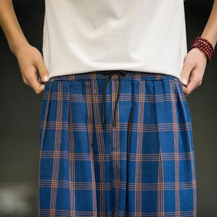 A person wearing a white shirt and blue plaid cotton joggers holds the waistband of the pants, making a subtle fashion statement. A beaded bracelet adorns their right wrist. The dark, out-of-focus background adds a stylish contrast.