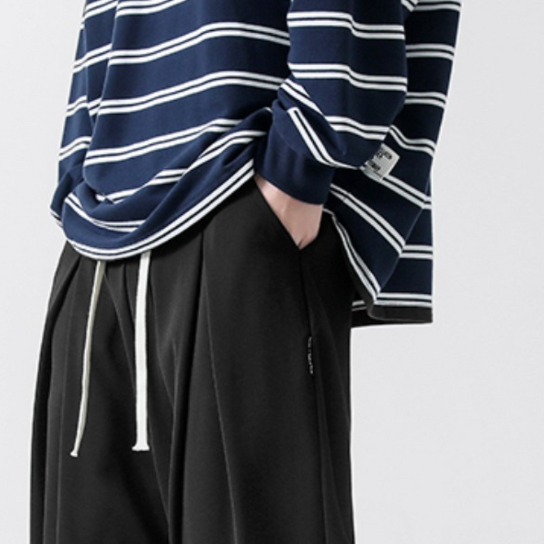 A person wears a navy blue and white striped long-sleeve shirt with loose black streetwear joggers featuring white drawstrings. One hand is in their pocket, and their head is not visible in the image.
