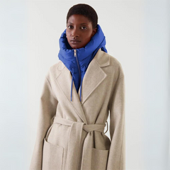 A person wearing a beige belted coat over a bright blue puffer jacket with a hooded design, standing against a plain light background.
