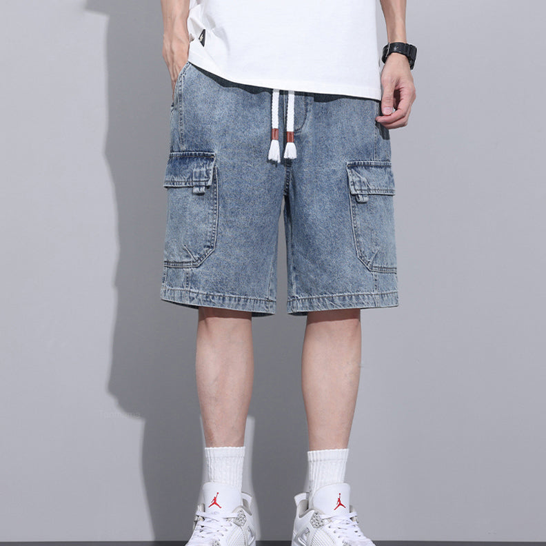 A person wearing light blue mens denim shorts with an elastic waist, a white T-shirt, white socks, and white sneakers stands against a gray background. Only the lower half of their body is visible.