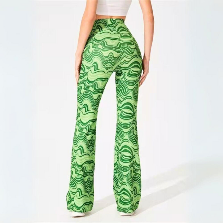 A person wearing high-waisted, psychedelic flare pants with a wavy green pattern stands facing away from the camera. They pair these trendy statement pants with a white crop top against a plain, light-colored background.