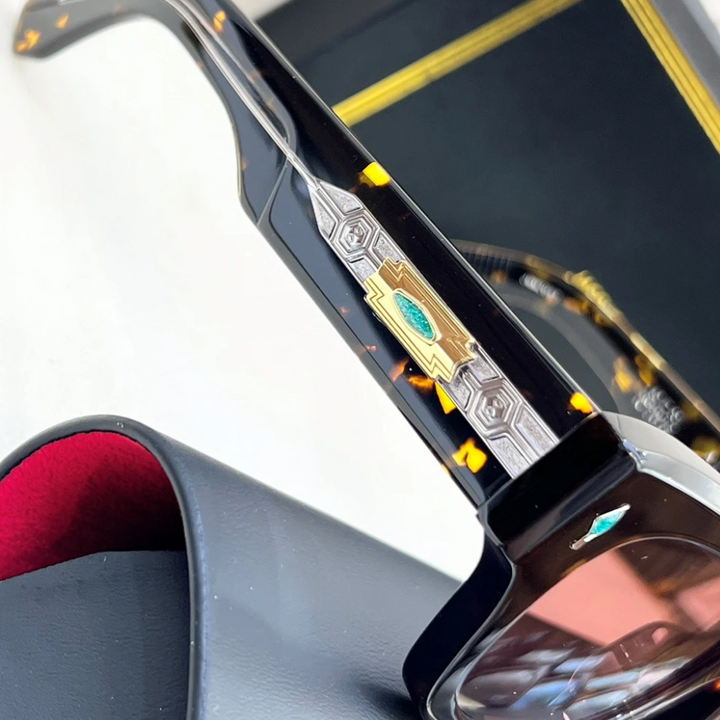 Close-up of retro sunglasses with tortoiseshell frames, silver and gold metal detailing, UV400 protection, and a green gem on the temple. Part of a black case with red lining and a black box with gold trim are also visible.