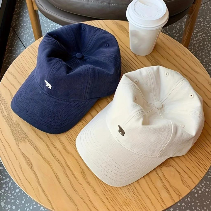 Two baseball caps, crafted from premium cotton—one navy blue and one off-white—lie on a light wooden table beside a white takeaway coffee cup. Each cap features a small, minimalist animal logo on the front, offering style and sun protection.