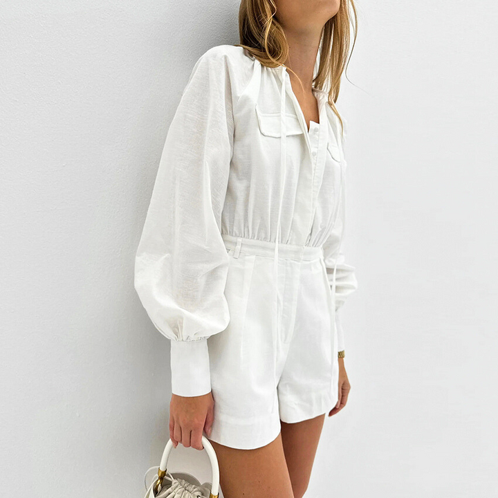 Woman wearing a chic romper with long sleeves, pockets, and a cinched waist, holding a small beige handbag—perfect streetwear style for a summer outing—standing against a plain white wall.