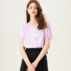 A young woman with long brown hair wears a light purple lace trim blouse with pearl-like buttons and a black high-waisted skirt, standing against a plain light background.