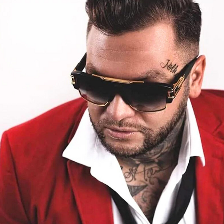 A man with slicked-back hair, sporting geometric sunglasses with scratch-resistant lenses and UV400 protection, a red blazer over a white shirt, loosely tied black tie, tattoos on his neck, and a script tattoo on his temple.
